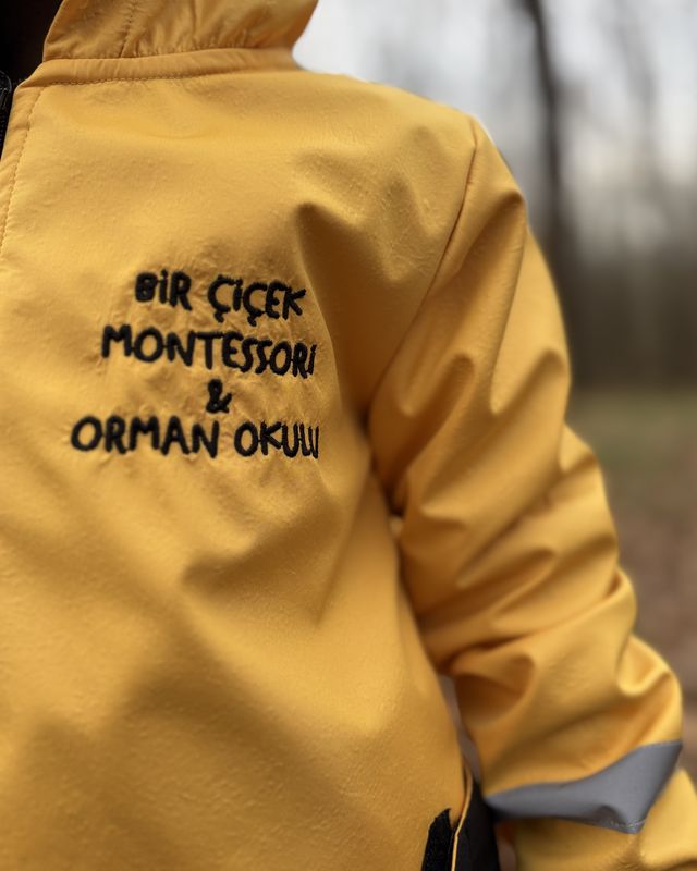 Orman okulunda sarı yağmurluk giymiş bir çocuk; montun üzerinde Bir Çiçek Montessori ve Orman Okulu yazıyor
